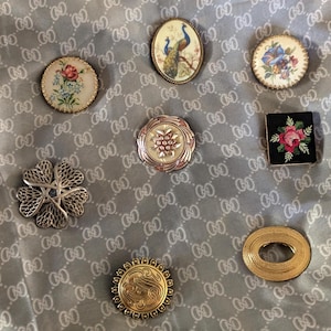Op de afbeelding: Een verzameling van acht diverse broches, waaronder ronde bloemmotieven, een ovaal pauwontwerp, een vierkant zwart stuk met een rode bloem, een zilverkleurige filigraanbloem, een geschulpt metalen stuk en een effen goudkleurige ovale broche.
