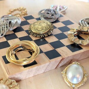 May include: A collection of assorted metallic and pearl-toned brooches arranged on a wooden board with a black and white checkerboard pattern. The pins feature various circular, oval, and abstract shapes in gold and silver tones with textured details.
