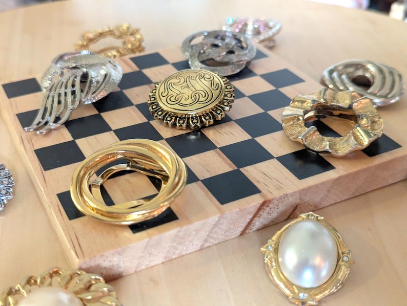 May include: A collection of assorted metallic and pearl-toned brooches arranged on a wooden board with a black and white checkerboard pattern. The pins feature various circular, oval, and abstract shapes in gold and silver tones with textured details.