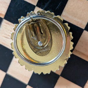 May include: A gold-toned circular clip-on earring with a scalloped edge and a central spring-loaded clip mechanism. The back of the earring features a stamped inscription that reads "GERMANY" in capital letters, indicating its origin.