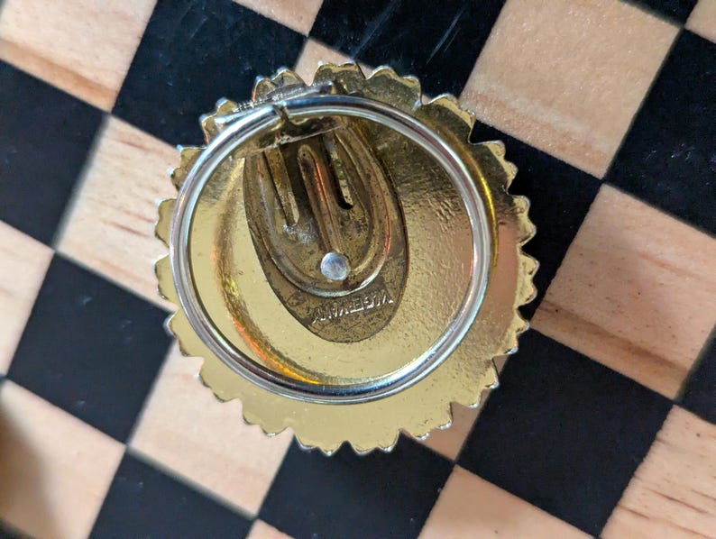 May include: A gold-toned circular clip-on earring with a scalloped edge and a central spring-loaded clip mechanism. The back of the earring features a stamped inscription that reads "GERMANY" in capital letters, indicating its origin.