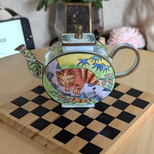 May include: A small, round, decorative metal teapot featuring a colorful enamel illustration of a brown tabby cat resting on a branch. The teapot has a brass-colored handle, spout, and lid knob, with green, yellow, and floral accents surrounding the cat.