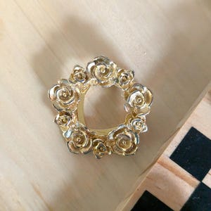 May include: A circular gold-toned metal brooch or scarf ring featuring a relief design of multiple blooming roses arranged in a wreath pattern. The metallic surface has a polished finish with detailed petal textures throughout the floral arrangement.