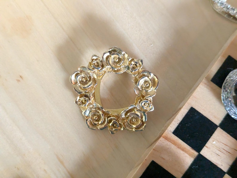 May include: A circular gold-toned metal brooch or scarf ring featuring a relief design of multiple blooming roses arranged in a wreath pattern. The metallic surface has a polished finish with detailed petal textures throughout the floral arrangement.