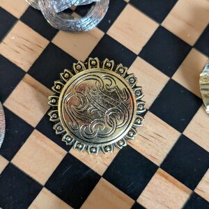 May include: A circular gold-toned metal button featuring an intricate embossed monogram design in the center, surrounded by a decorative scalloped border with small raised dots. The button has a polished finish and a detailed, ornate appearance.