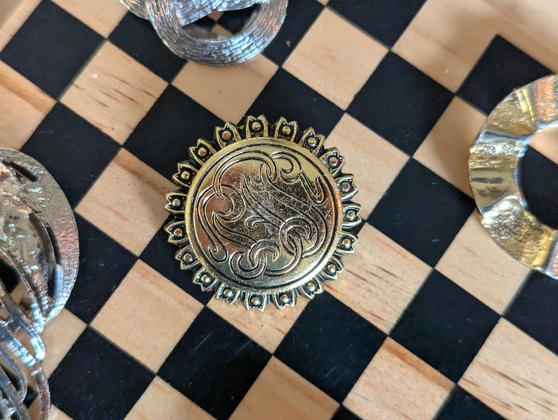 May include: A circular gold-toned metal button featuring an intricate embossed monogram design in the center, surrounded by a decorative scalloped border with small raised dots. The button has a polished finish and a detailed, ornate appearance.