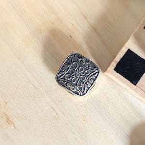 May include: A square-shaped metal button featuring a raised, intricate floral pattern with a dark, oxidized finish in the recessed areas and a polished, metallic finish on the elevated surfaces, showcasing a symmetrical, repeating design.