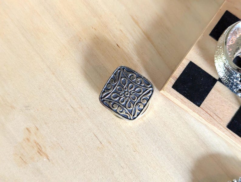 May include: A square-shaped metal button featuring a raised, intricate floral pattern with a dark, oxidized finish in the recessed areas and a polished, metallic finish on the elevated surfaces, showcasing a symmetrical, repeating design.