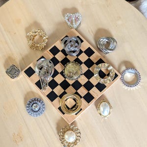 May include: A collection of twelve assorted metal brooches and pins featuring various shapes, including circular, oval, and heart designs. The pieces display gold and silver tones, some with embedded faux pearls, clear crystals, and intricate textures.