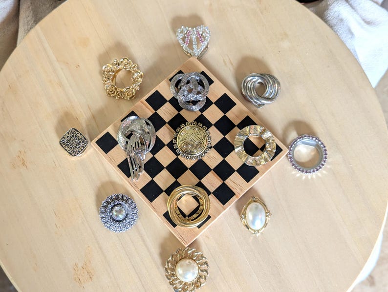 May include: A collection of twelve assorted metal brooches and pins featuring various shapes, including circular, oval, and heart designs. The pieces display gold and silver tones, some with embedded faux pearls, clear crystals, and intricate textures.