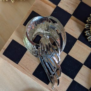 May include: A silver-toned metal brooch featuring a circular frame with a textured finish and a central cascading element composed of multiple curved, elongated strips that taper toward the bottom, creating a fluid and layered appearance.