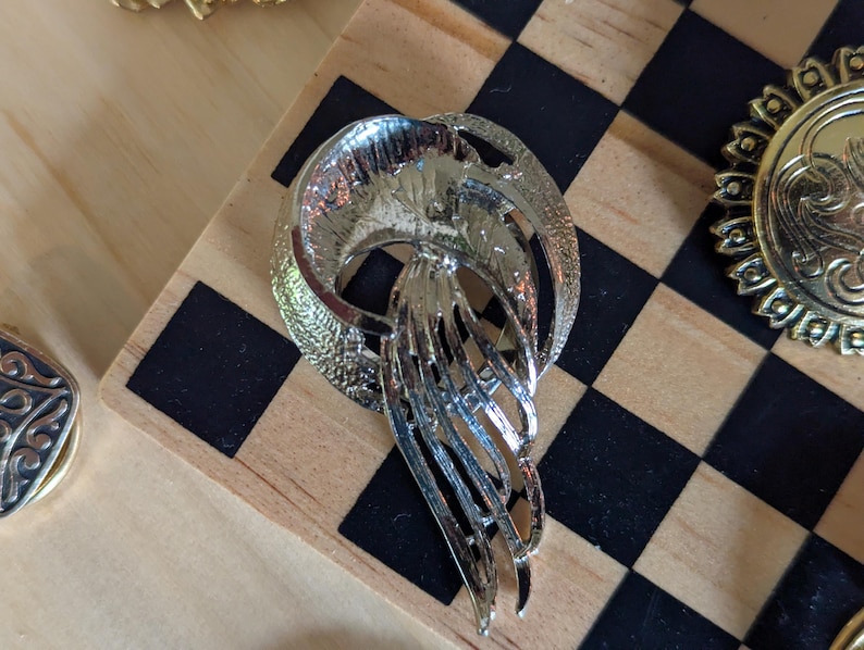 May include: A silver-toned metal brooch featuring a circular frame with a textured finish and a central cascading element composed of multiple curved, elongated strips that taper toward the bottom, creating a fluid and layered appearance.