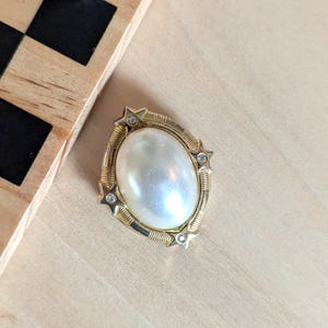 May include: An oval brooch featuring a central white faux pearl cabochon set in a gold-tone metal frame. The frame is decorated with four small star-shaped accents, each containing a tiny clear rhinestone, and textured ribbed metal detailing.