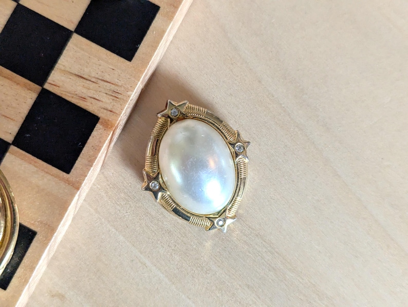 May include: An oval brooch featuring a central white faux pearl cabochon set in a gold-tone metal frame. The frame is decorated with four small star-shaped accents, each containing a tiny clear rhinestone, and textured ribbed metal detailing.