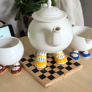 Puede incluir: Un juego de té de cerámica que consta de una tetera redonda, una jarra de leche y un azucarero, cada uno con patas antropomórficas con zapatos pintados. La tetera se apoya sobre una tabla de madera cuadrada con un patrón de tablero de ajedrez.