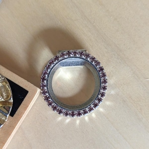 May include: A circular silver-toned ring featuring a band encrusted with small, light purple faceted stones. The inner surface of the ring has a textured, grid-like pattern. A small rectangular metal tab extends from the top edge of the ring.