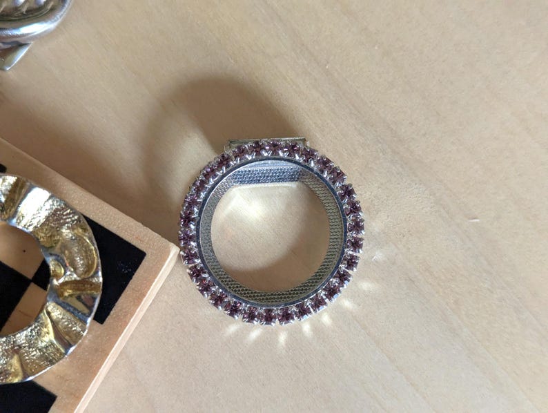 May include: A circular silver-toned ring featuring a band encrusted with small, light purple faceted stones. The inner surface of the ring has a textured, grid-like pattern. A small rectangular metal tab extends from the top edge of the ring.