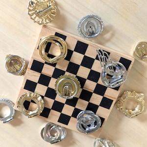 May include: A collection of various gold and silver tone metal clip-on earrings arranged around a small square wooden board featuring a black and white checkered pattern. The earrings display diverse circular, textured, and wing-like shapes.