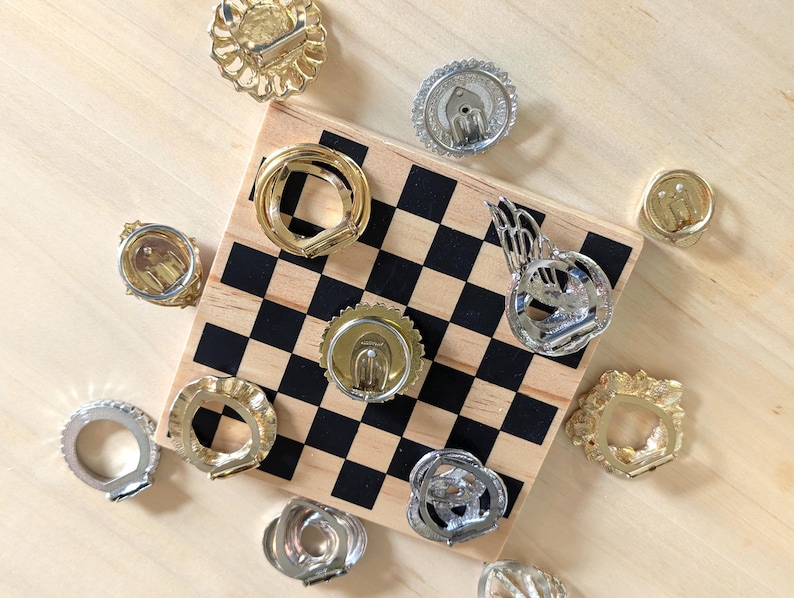 May include: A collection of various gold and silver tone metal clip-on earrings arranged around a small square wooden board featuring a black and white checkered pattern. The earrings display diverse circular, textured, and wing-like shapes.
