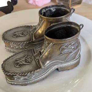 May include: A pair of metallic, silver-toned decorative boots featuring embossed floral motifs on the toe and side panels. The boots have a textured sole, a defined heel, and a small loop at the back of the collar, with a tarnished finish.