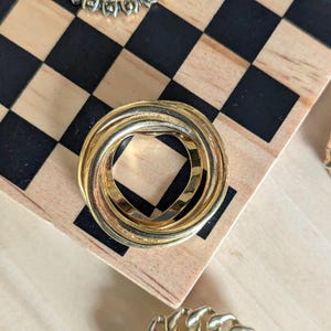 May include: A circular brooch composed of three interlocking gold-toned metal bands. The design features a smooth, polished finish on the outer rings and a textured, matte finish on the central band, creating a layered, continuous loop appearance.