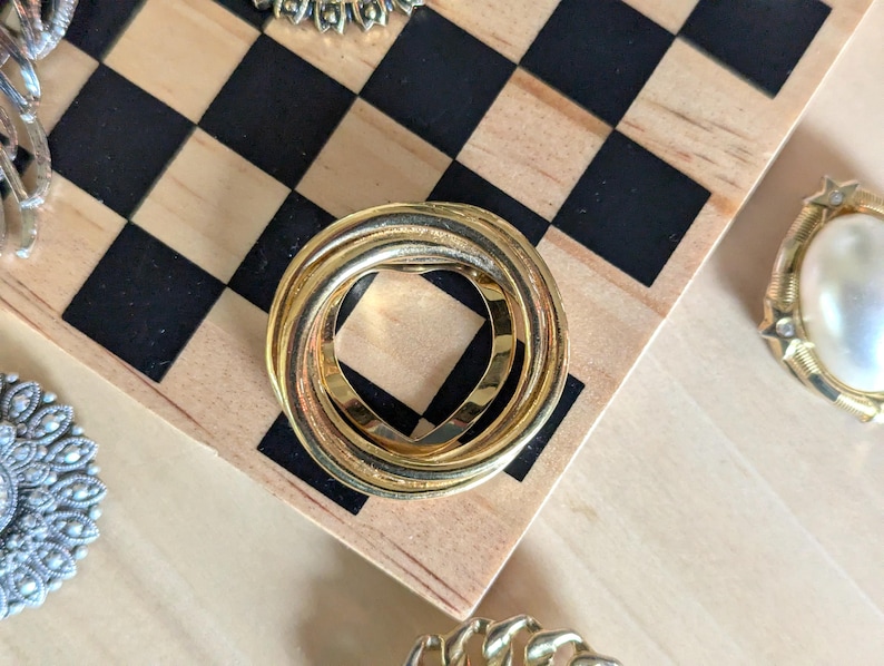 May include: A circular brooch composed of three interlocking gold-toned metal bands. The design features a smooth, polished finish on the outer rings and a textured, matte finish on the central band, creating a layered, continuous loop appearance.