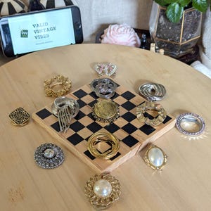 May include: A collection of eleven assorted metal scarf rings and brooches in gold and silver tones, featuring various textures, filigree patterns, and faux pearl accents, arranged on a small wooden square board with a black and white checkerboard pattern.