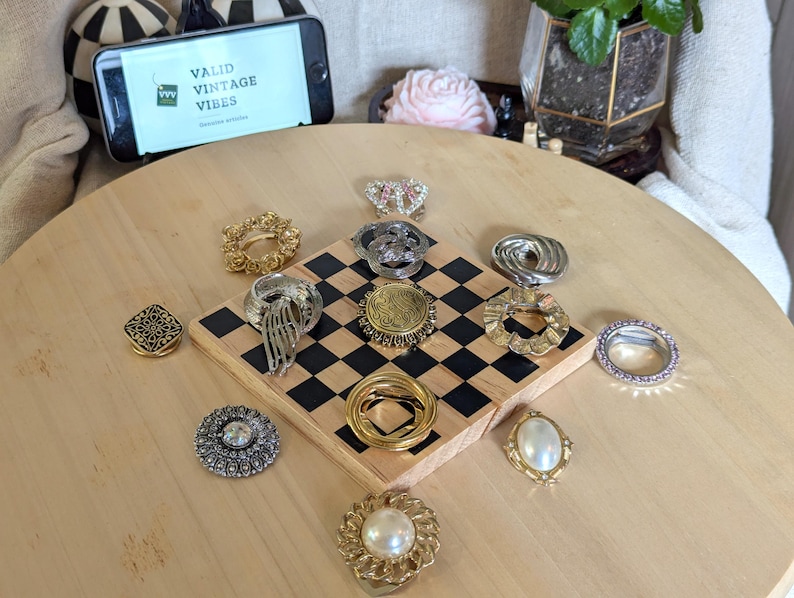 May include: A collection of eleven assorted metal scarf rings and brooches in gold and silver tones, featuring various textures, filigree patterns, and faux pearl accents, arranged on a small wooden square board with a black and white checkerboard pattern.