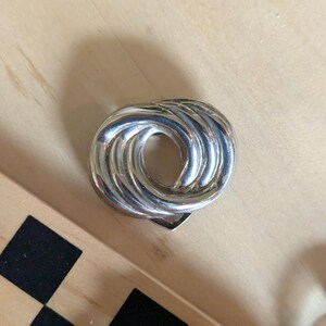 May include: A polished silver-tone metal brooch featuring a circular, knotted design with multiple overlapping loops. The piece has a reflective, smooth surface with a central opening and a secure pin mechanism visible on the back side.