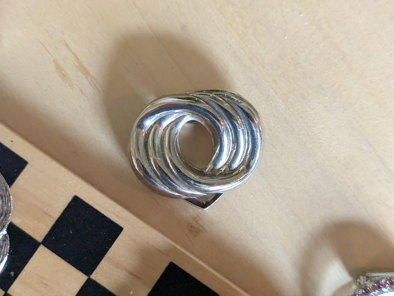 May include: A polished silver-tone metal brooch featuring a circular, knotted design with multiple overlapping loops. The piece has a reflective, smooth surface with a central opening and a secure pin mechanism visible on the back side.