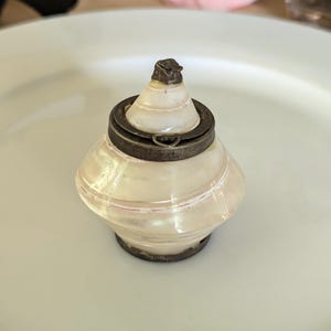 Op de afbeelding: Een klein, parelmoerkleurig, kegelvormig object met een glad, wit en crèmekleurig wervelend oppervlak. Het is voorzien van een donkere, verweerde metalen band rond het midden en een bijpassende metalen voet, bekroond met een kleine, puntige metalen pit.