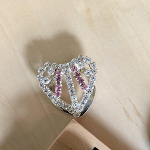 May include: A heart-shaped brooch featuring a silver-toned metal frame encrusted with clear white rhinestones and two vertical rows of small pink rhinestones. The design includes an open center and a polished metallic base visible beneath the stones.