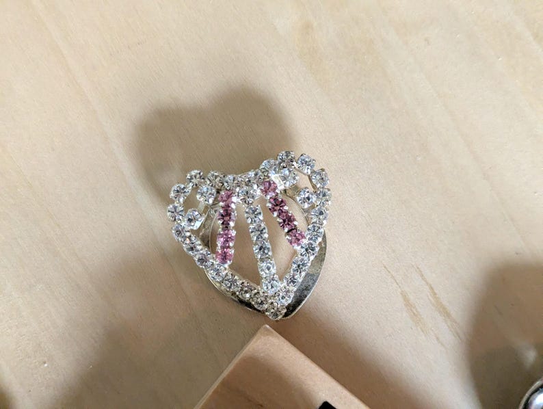 May include: A heart-shaped brooch featuring a silver-toned metal frame encrusted with clear white rhinestones and two vertical rows of small pink rhinestones. The design includes an open center and a polished metallic base visible beneath the stones.