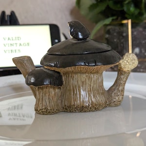 Puede incluir: Una tetera de cerámica con forma de grupo de setas con un sombrero marrón oscuro y un tallo beige texturizado. La tetera cuenta con una pequeña tapa con forma de seta y un asa curva en el lateral. El diseño incluye detalles realistas.