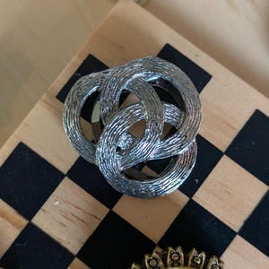 May include: A silver-toned metal brooch featuring three interlocking circular rings with a textured, brushed finish. The design consists of three overlapping loops that create a continuous, knotted appearance, showcasing a metallic sheen throughout.