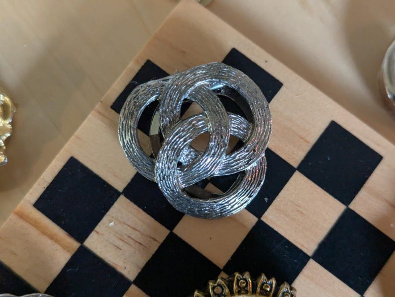 May include: A silver-toned metal brooch featuring three interlocking circular rings with a textured, brushed finish. The design consists of three overlapping loops that create a continuous, knotted appearance, showcasing a metallic sheen throughout.