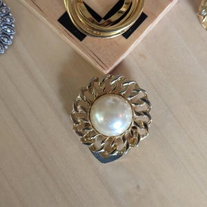 May include: A circular brooch featuring a central, smooth, cream-colored faux pearl surrounded by a textured, gold-tone metal frame with an interlocking chain-link design. The piece has a polished finish and a round, symmetrical structure.