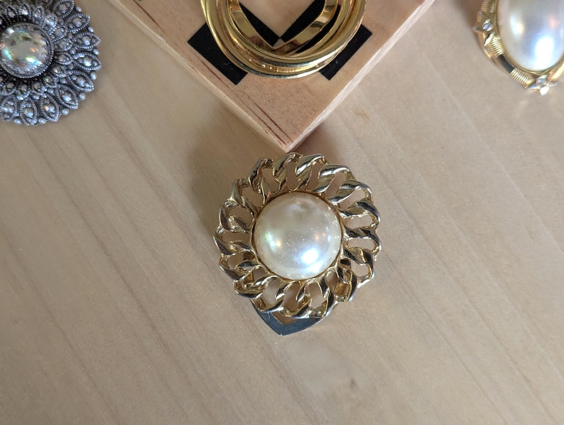 May include: A circular brooch featuring a central, smooth, cream-colored faux pearl surrounded by a textured, gold-tone metal frame with an interlocking chain-link design. The piece has a polished finish and a round, symmetrical structure.