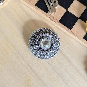 May include: A circular silver-toned brooch featuring an intricate filigree pattern radiating from a central iridescent cabochon stone. The metalwork displays a dark oxidized finish within the recessed areas, highlighting the detailed floral-like design.