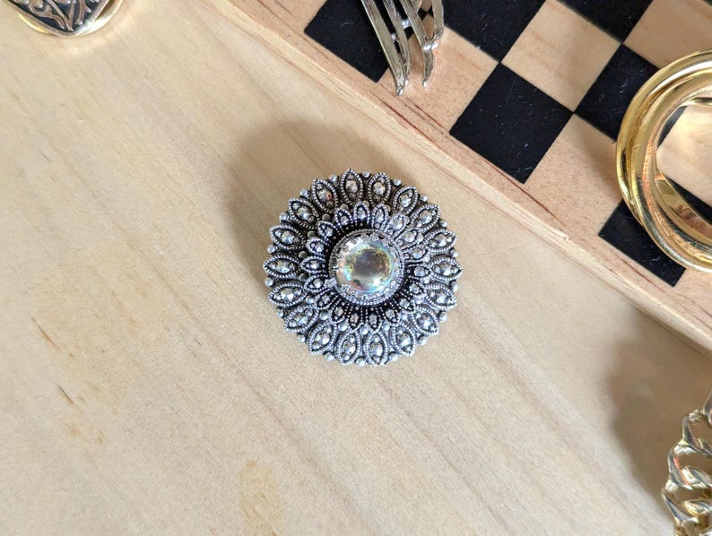 May include: A circular silver-toned brooch featuring an intricate filigree pattern radiating from a central iridescent cabochon stone. The metalwork displays a dark oxidized finish within the recessed areas, highlighting the detailed floral-like design.