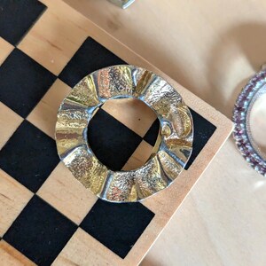 May include: A circular metal brooch with a wavy, rippled edge and a textured, gold-toned finish. The center is open, creating a ring-like shape. The surface features alternating smooth and rough patches that catch the light, giving it a reflective appearance.