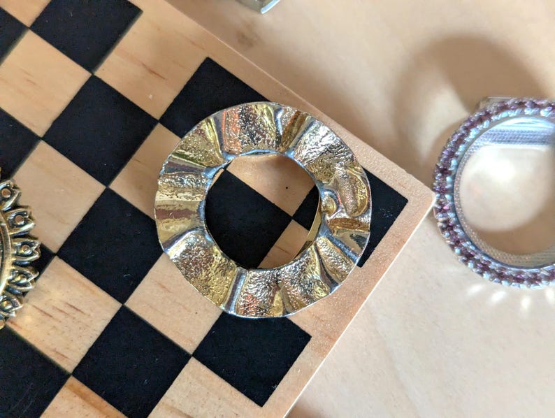 May include: A circular metal brooch with a wavy, rippled edge and a textured, gold-toned finish. The center is open, creating a ring-like shape. The surface features alternating smooth and rough patches that catch the light, giving it a reflective appearance.