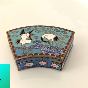 May include: A fan-shaped metal box with cloisonné enamel featuring two white cranes on a light blue background with cloud motifs. The sides of the box are decorated with pink floral patterns on a blue field, framed by thin gold-toned metal wire.