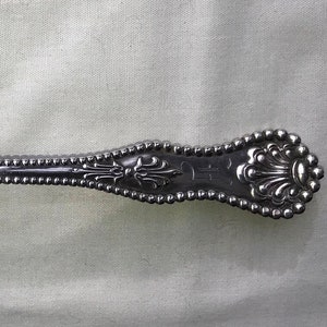 Puede incluir: Una cuchara de plata con un cuenco ovalado y un mango decorativo. El mango presenta un diseño de cuentas y detalles florales ornamentados. La cuchara es probablemente de plata de ley y es una pieza de cubertería vintage.