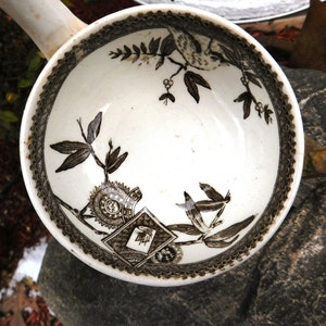 Könnte beinhalten: Eine weiße Keramikschale mit braunem Blumen- und Vogelmuster. Der Rand hat eine dekorative Bordüre. Die Schale hat einen Griff. Das Design umfasst Blätter, einen Vogel und geometrische Formen.