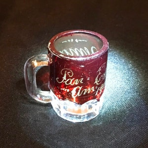 Op de afbeelding: Een klein, helder glazen mok met een rode, ondoorzichtige glazen inzet. De mok heeft een handvat en de woorden "Pan-Am" en "Am's" zijn gegraveerd in de inzet.