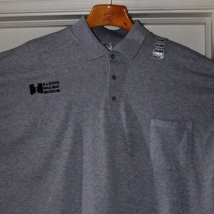 Puede incluir: Una camiseta polo gris de manga corta con un bolsillo en el lado izquierdo. La camiseta tiene un logotipo negro que dice "Baltimore Railway Museum".