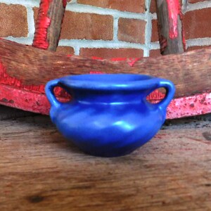 以下が含まれることがあります： 2つの取っ手が付いた小さな青い陶器の壺。壺は木製の表面の上に置かれています。