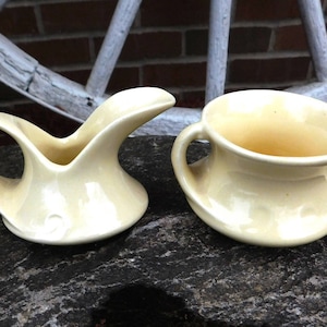 Könnte beinhalten: Ein Set aus zwei vintage Keramik-Sahnegießern und Zuckerbehältern in blassgelber Farbe. Der Sahnegießer hat eine einzigartige Form mit einem gebogenen Ausguss und Griff. Der Zuckerbehälter hat zwei Griffe und eine abgerundete Form.