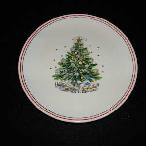 May include: White ceramic plate with a Christmas tree design. The tree is decorated with ornaments and topped with a star. A red and blue stripe border frames the plate, and a village scene is at the base of the tree.